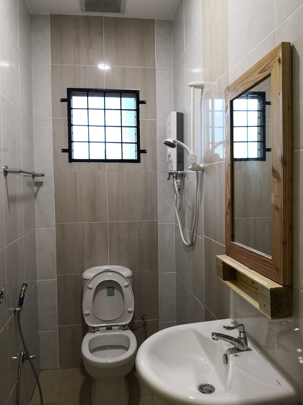 A compact bathroom features light-colored tiles, a modern toilet, and a wall-mounted sink with a single faucet. A showerhead is visible, alongside a wooden-framed mirror mounted on the wall. A small window allows natural light to enter, covered by a grid design.