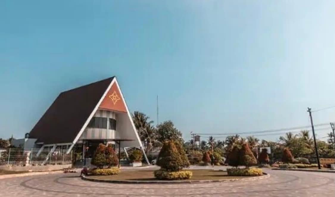 A modern villa features a distinct A-frame architecture with a triangular roof and decorative detailing. Surrounding greenery includes well-trimmed shrubs and small trees, enhancing the warm exterior. A circular driveway offers easy access, and a clear blue sky creates a serene backdrop.