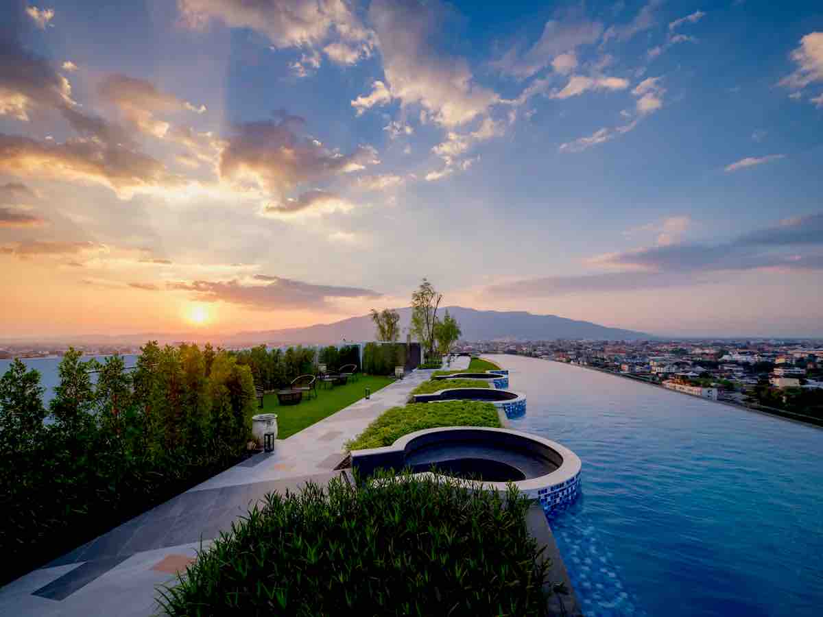 The rooftop area features an expansive infinity pool with a stunning view of the sunset and surrounding mountains. Lush greenery surrounds the pool, complemented by stylish seating areas. The atmosphere conveys a sense of tranquility and relaxation, perfect for unwinding after a day in the city.