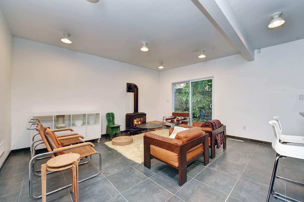 A spacious living area showcases modern furnishings, including a comfortable wooden sofa and distinctive chairs. Natural light enters through glass doors that lead to a garden. A minimalist fireplace complements the white walls and large tiles, inviting a cozy atmosphere.