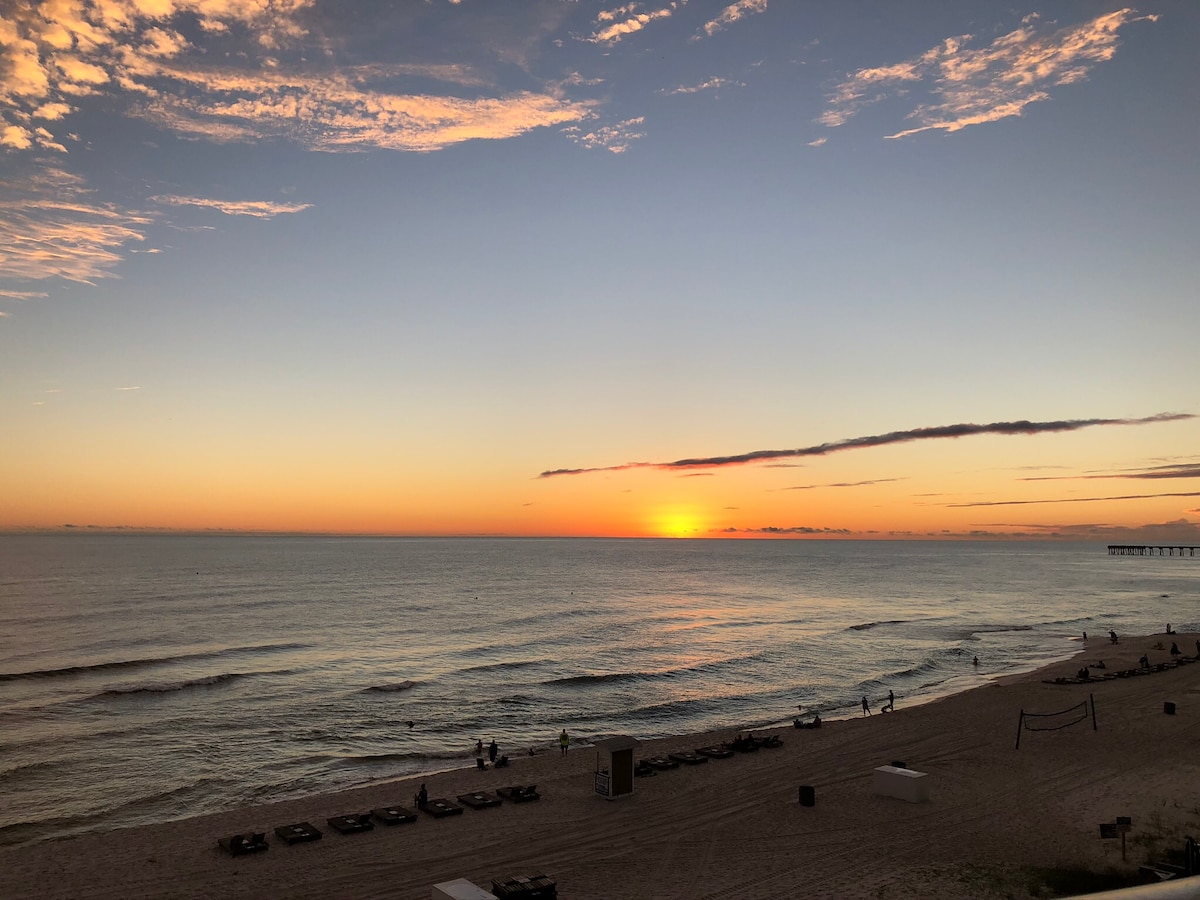 A serene beachfront scene captures the ocean at sunset. Gentle waves roll onto the shore, reflecting warm hues of orange and pink in the sky. Sun loungers are arranged on the sand, and silhouettes of beachgoers can be seen in the distance enjoying the tranquil atmosphere.