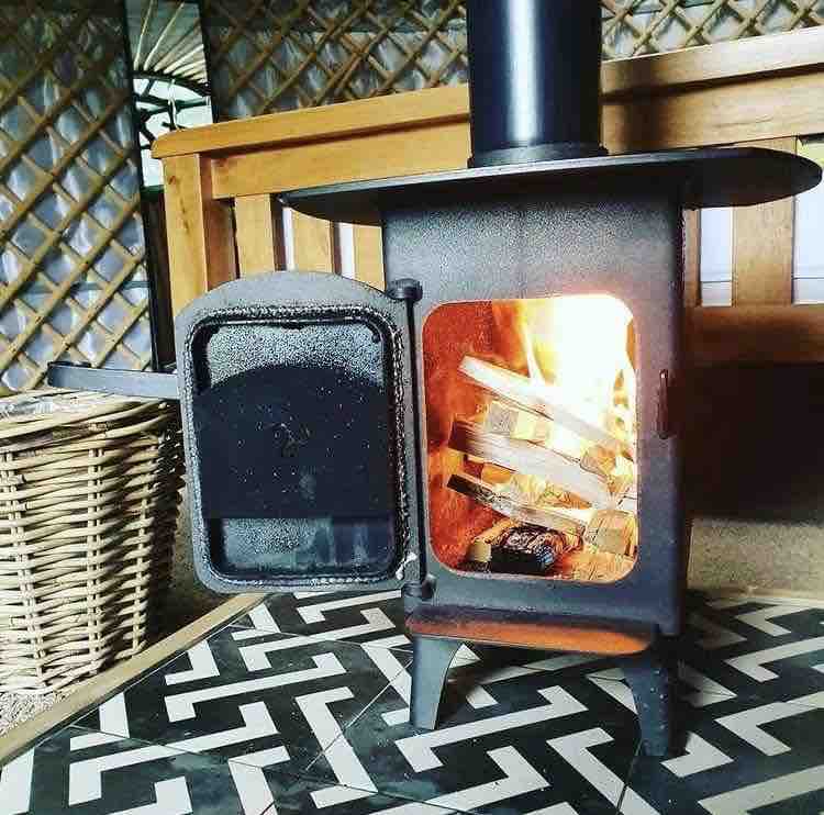 A pleasing wood-burning stove is positioned inside the yurt, featuring a black metal body and an open door revealing gently flickering flames and burning logs. The stove sits on a patterned tile floor, complemented by a woven basket nearby.