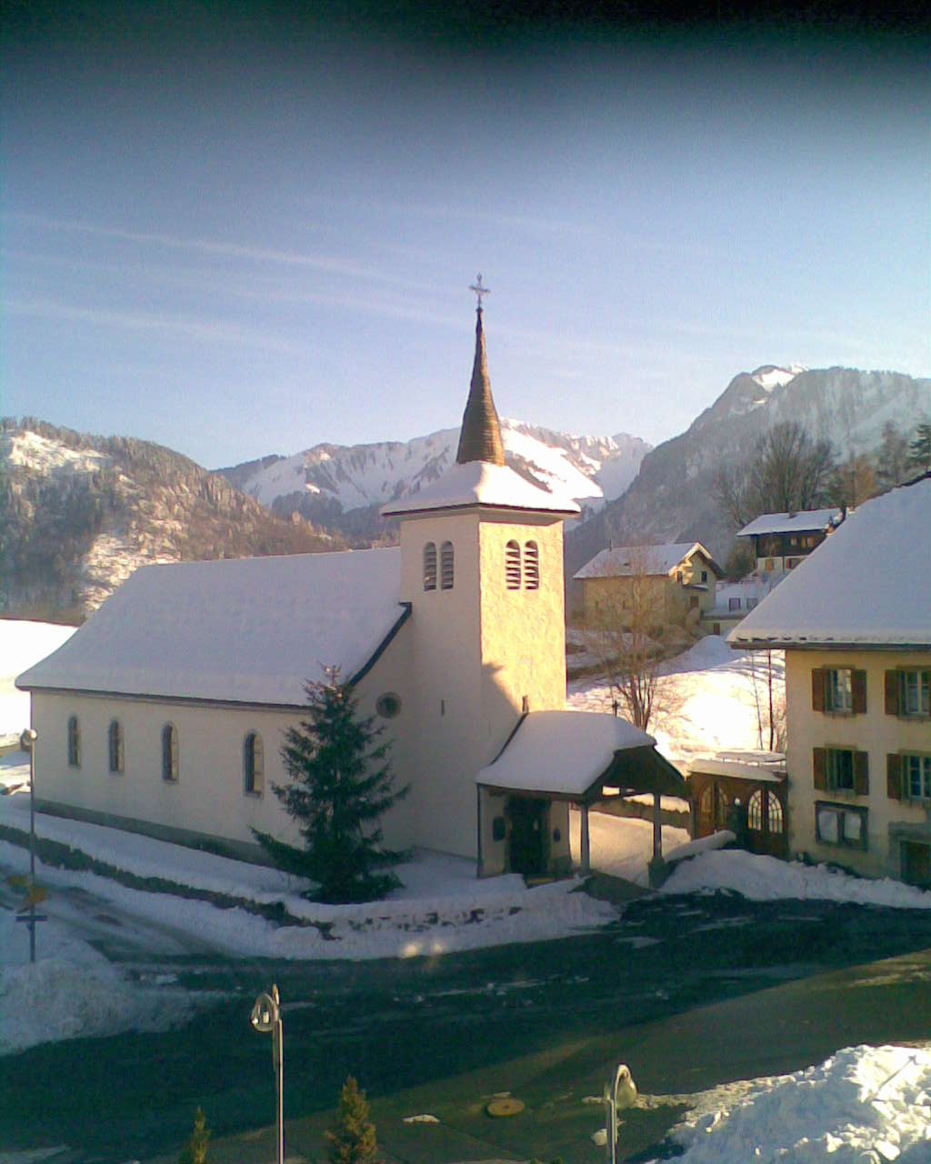 A quaint church is depicted, covered in a blanket of snow, with a prominent steeple reaching towards the sky. Snow-laden mountains surround the scene, and charming buildings are visible in the foreground, creating a serene winter landscape.