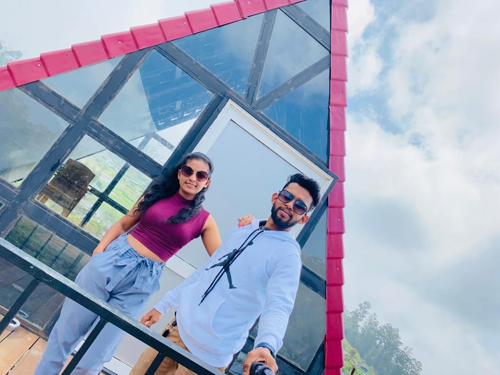 A modern structure with a distinctive glass facade and a red roof is depicted. A spacious balcony adorned with a railing provides expansive views of the surroundings. The scene captures two individuals standing together, enjoying the serene atmosphere.