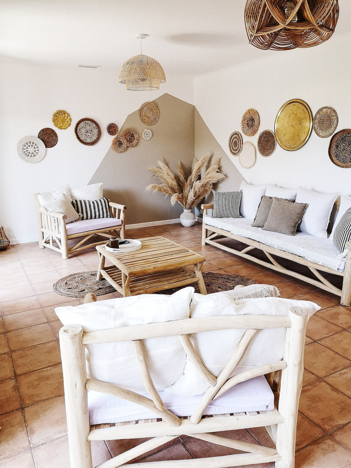 A welcoming living area features two light-toned wooden sofas adorned with soft cushions. A low wooden coffee table is centered on a woven rug. The walls display an assortment of decorative plates and natural elements, while a pendant light adds a soft glow to the space.