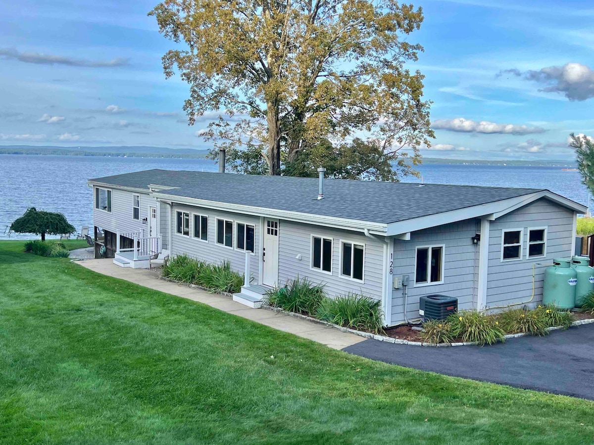 A single-story house with a light gray exterior is positioned on a lush green lawn. Large windows offer views of the lake, surrounded by trees. A paved path leads to the entrance, while the serene water and sky create a peaceful background.