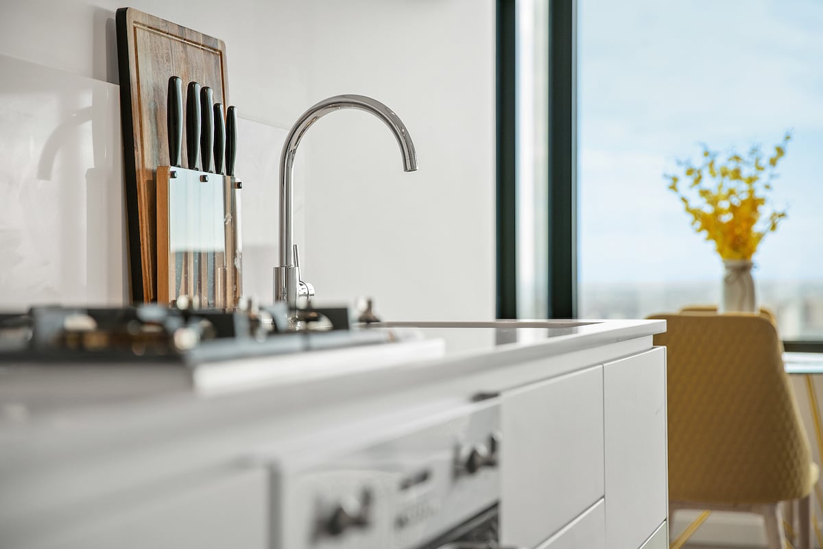 The kitchen area features sleek cabinetry with a modern sink and faucet. A wooden cutting board displays a set of knives below a window that allows natural light to illuminate the space. In the background, a table is set with a vase containing yellow flowers.