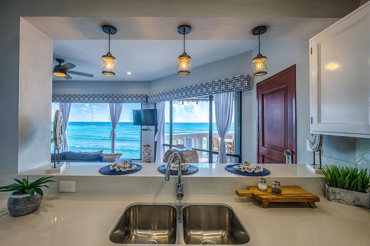 The kitchen area features a spacious countertop with a stainless steel sink, showcasing decorative plates and a wooden tray. Pendant lights hang above, illuminating the space. Large windows allow natural light to fill the room, revealing stunning ocean views just beyond the glass doors.
