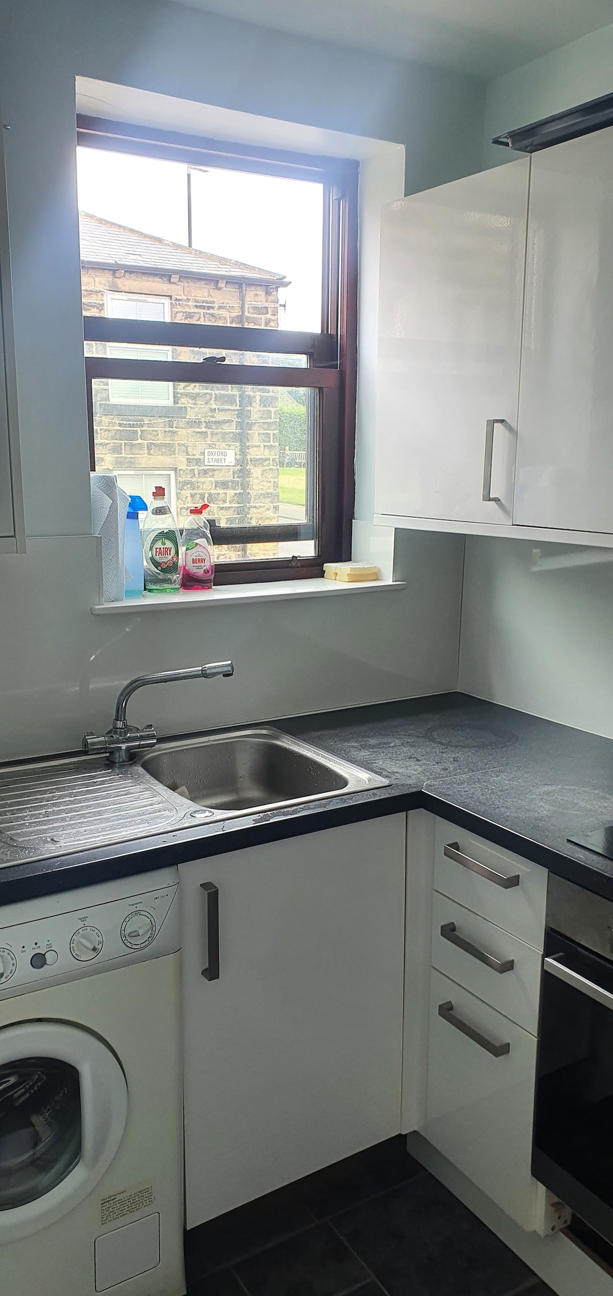 The kitchenette features a compact layout with white cabinetry and a dark countertop. A window provides natural light, showcasing nearby buildings. Appliances include a sink, stove, and washing machine, along with cleaning supplies displayed on the sill.