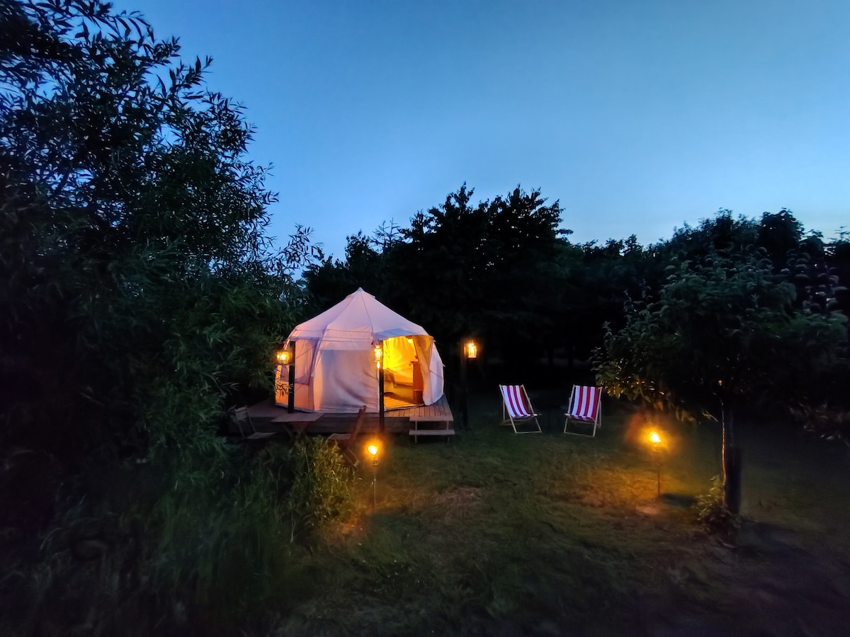 A spacious white tent is set against a twilight sky, emanating a warm glow from within. Two striped lounge chairs are positioned nearby, with soft lighting provided by lanterns and ground lights. Surrounding greenery adds a serene touch to the tranquil environment.