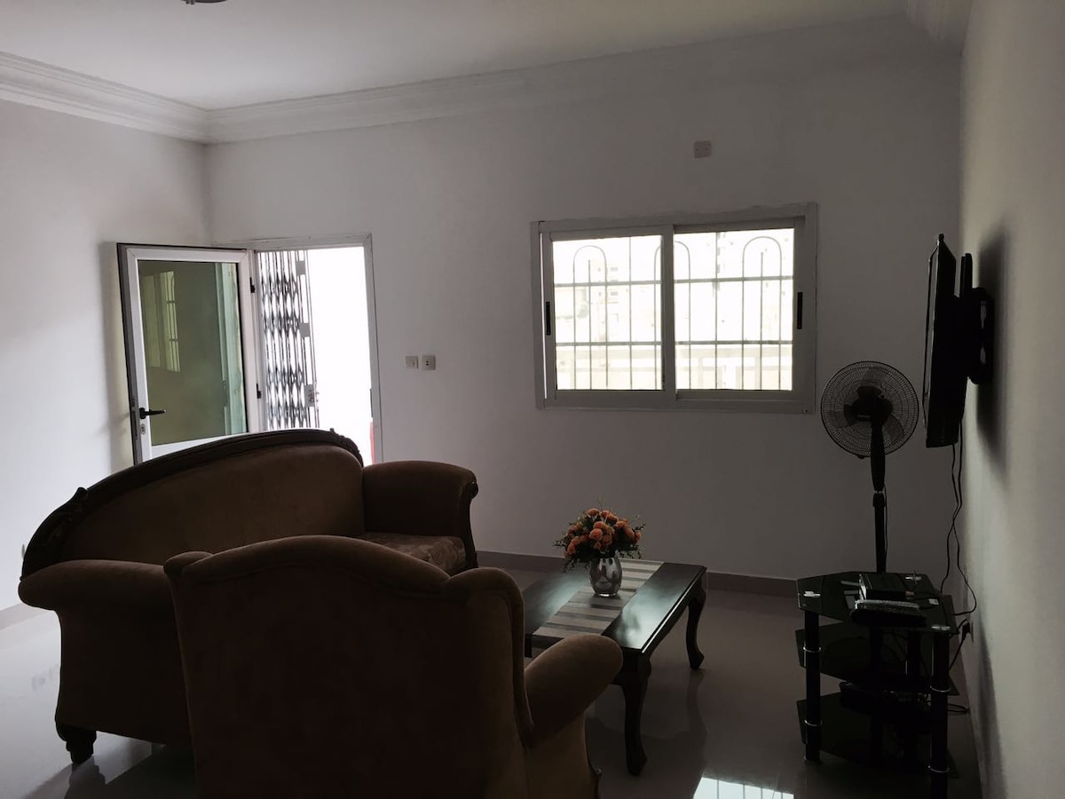 A living area is depicted, featuring a comfortable seating arrangement with a pair of armchairs and a wooden coffee table. A small vase of flowers rests on the table, while a television is mounted on a stand against the wall. Large windows allow natural light to enter the space.