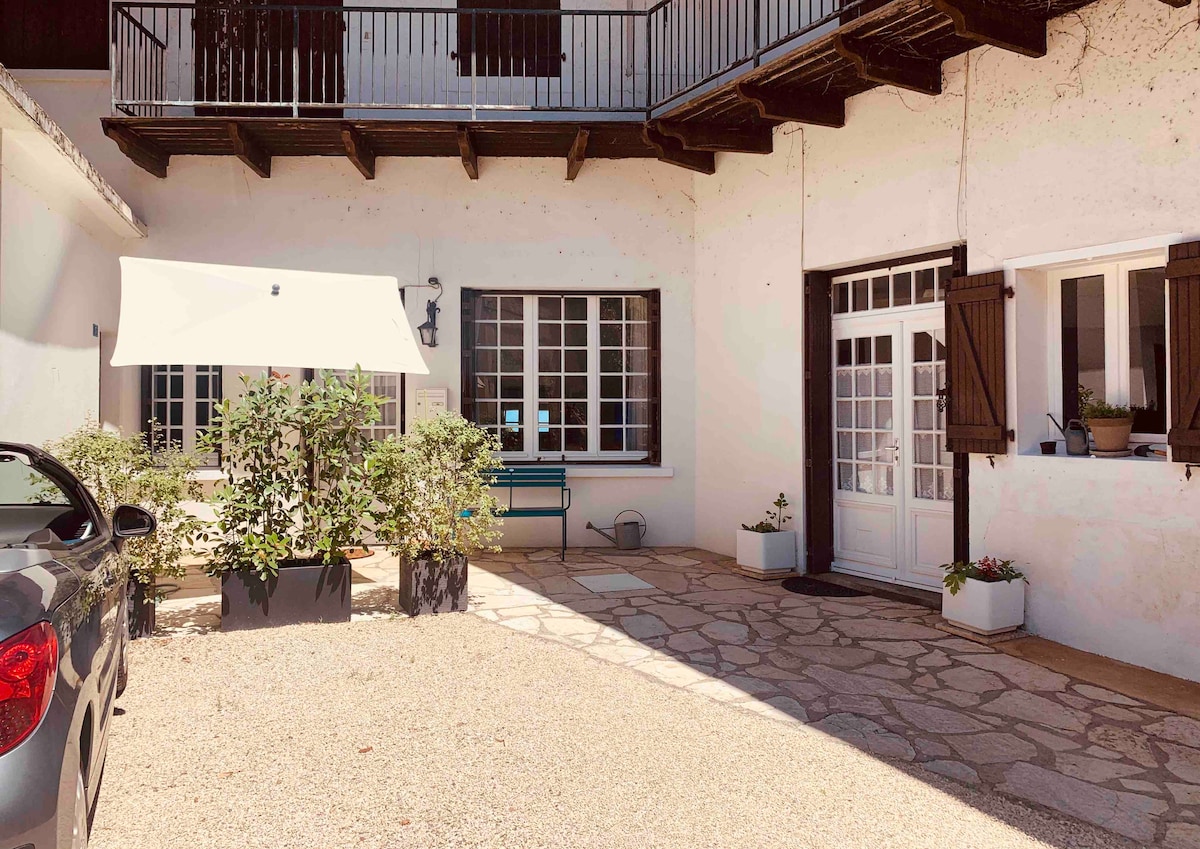 A charming outdoor courtyard is visible, featuring a stone patio, a retractable awning offering shade, and potted plants adding greenery. The entrance to the apartment has French doors with shutters, and a parked car is nearby, indicating convenient access.