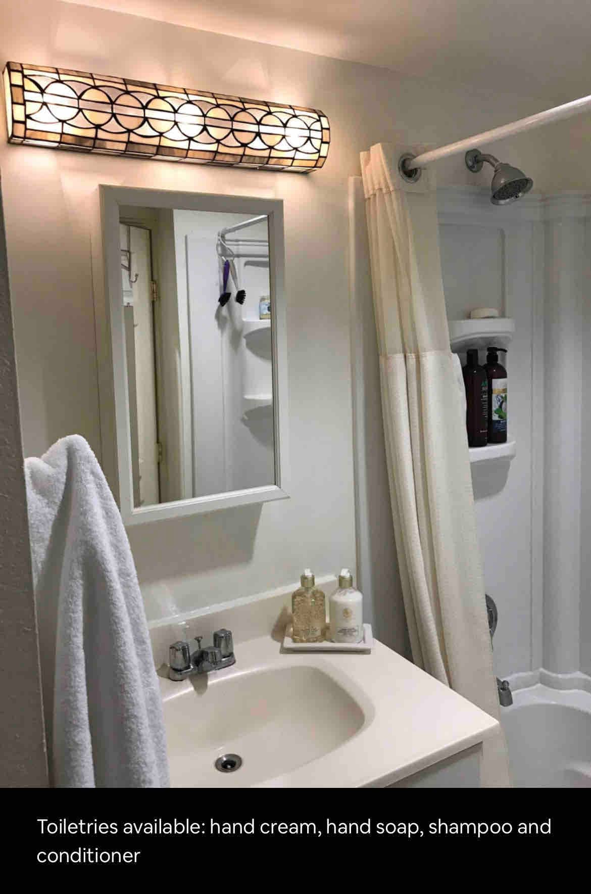 A clean bathroom is depicted, featuring a sink with a mirror above it. Toiletries including hand cream, hand soap, shampoo, and conditioner are organized on the countertop and in the shower area. A white towel hangs neatly by the sink.