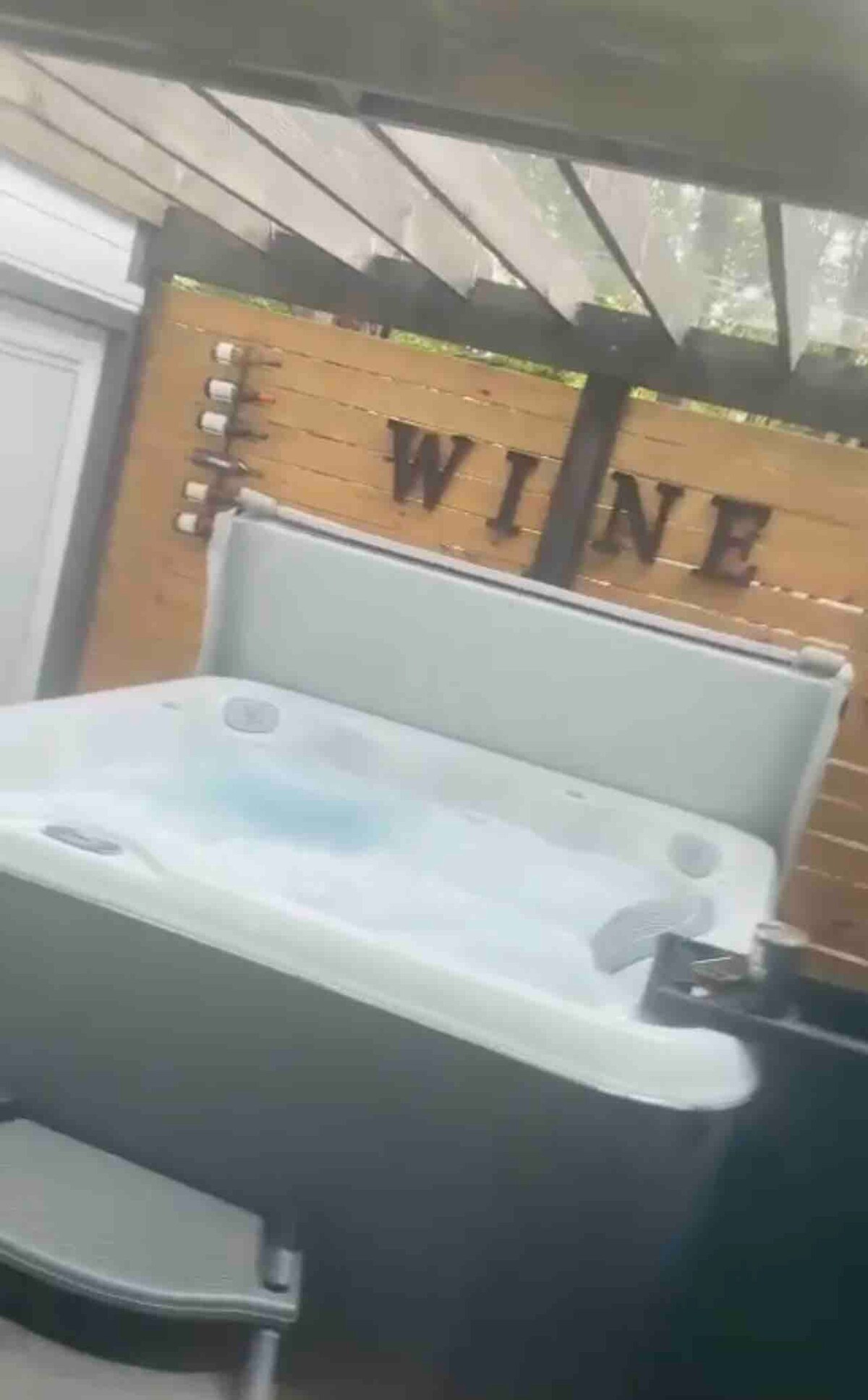 A hot tub is nestled in a serene outdoor area, featuring a wooden backdrop with the word 'WINE' displayed prominently. The hot tub is equipped with jets and steps leading to it, surrounded by a tranquil environment.