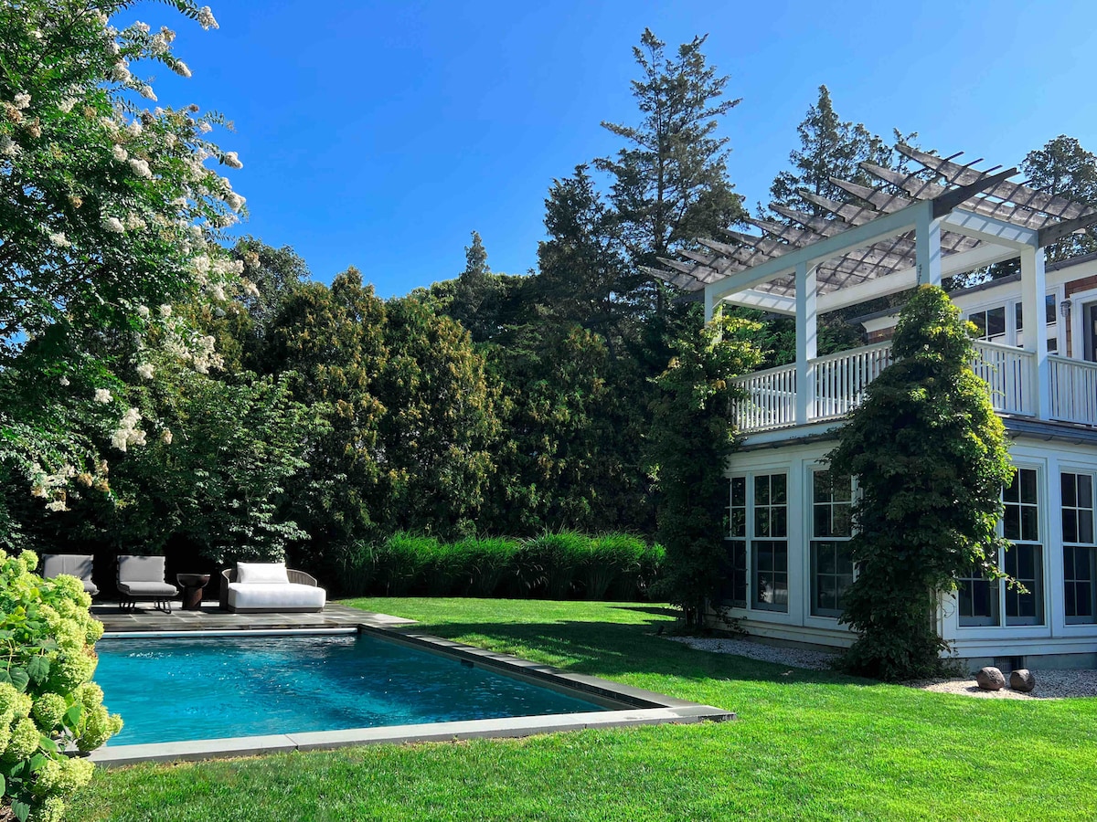 A glimmering gunite pool lies surrounded by lush greenery and a manicured lawn. The white house features a charming pergola overhead, with large windows offering a glimpse into the inviting interior. Sunny skies enhance the serene outdoor ambiance.