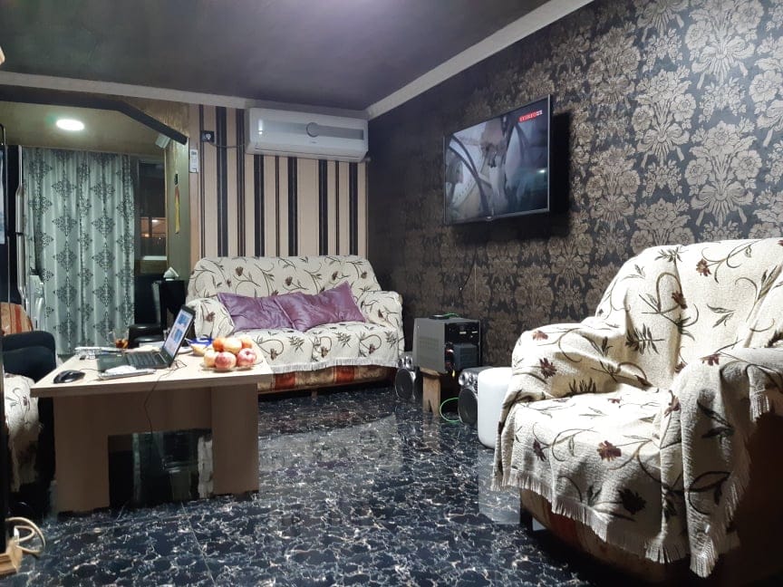A living room is presented with two upholstered sofas, both covered in patterned throws. A stylish coffee table sits at the center, adorned with fruit. A wall-mounted television displays a scene, while an air conditioning unit is visible above. The floor is finished in dark marbled tiles.