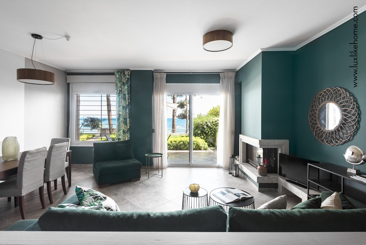 A spacious living area is filled with natural light, featuring a comfortable sectional sofa in rich green. A glass door leads to a garden, framed by curtains with a floral pattern. A modern fireplace and sleek black media console complement the design.