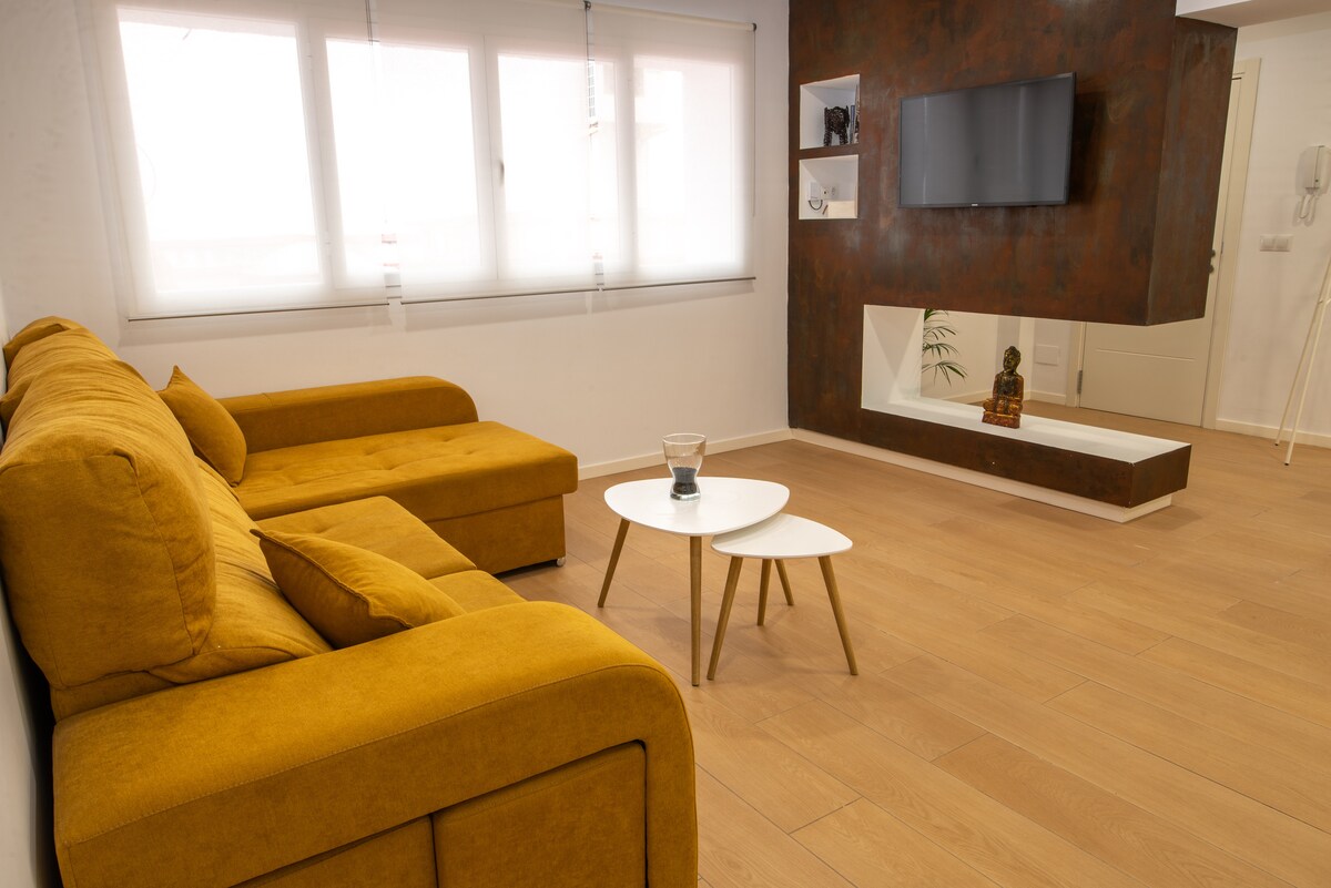 A modern living area features a mustard-colored sectional sofa paired with a round coffee table on light wooden flooring. A wall-mounted television is visible above a minimalist console, while large windows allow natural light to brighten the space.