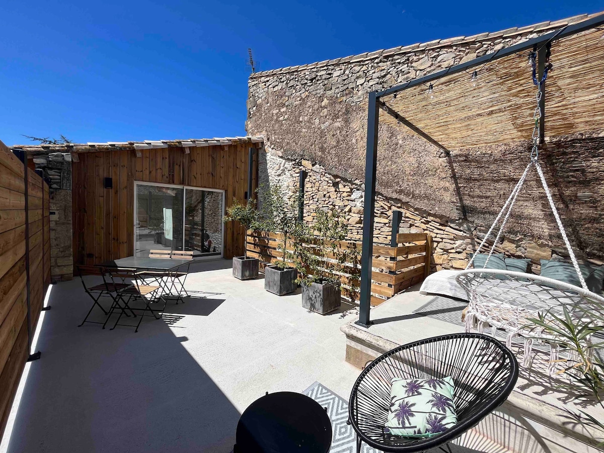 An outdoor terrace features various seating options, including a swing chair and two black lounge chairs. A wooden privacy fence creates an enclosed space, while natural stone walls add texture. Sunlight highlights the clean lines and modern design elements, contributing to a relaxing ambiance.