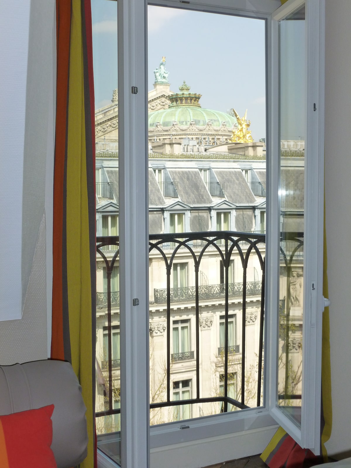 A view from the small balcony showcases the ornate rooftop of the Opéra, featuring a green dome topped with a golden figure. Brightly colored curtains frame the twin glass doors, allowing natural light to illuminate the space.