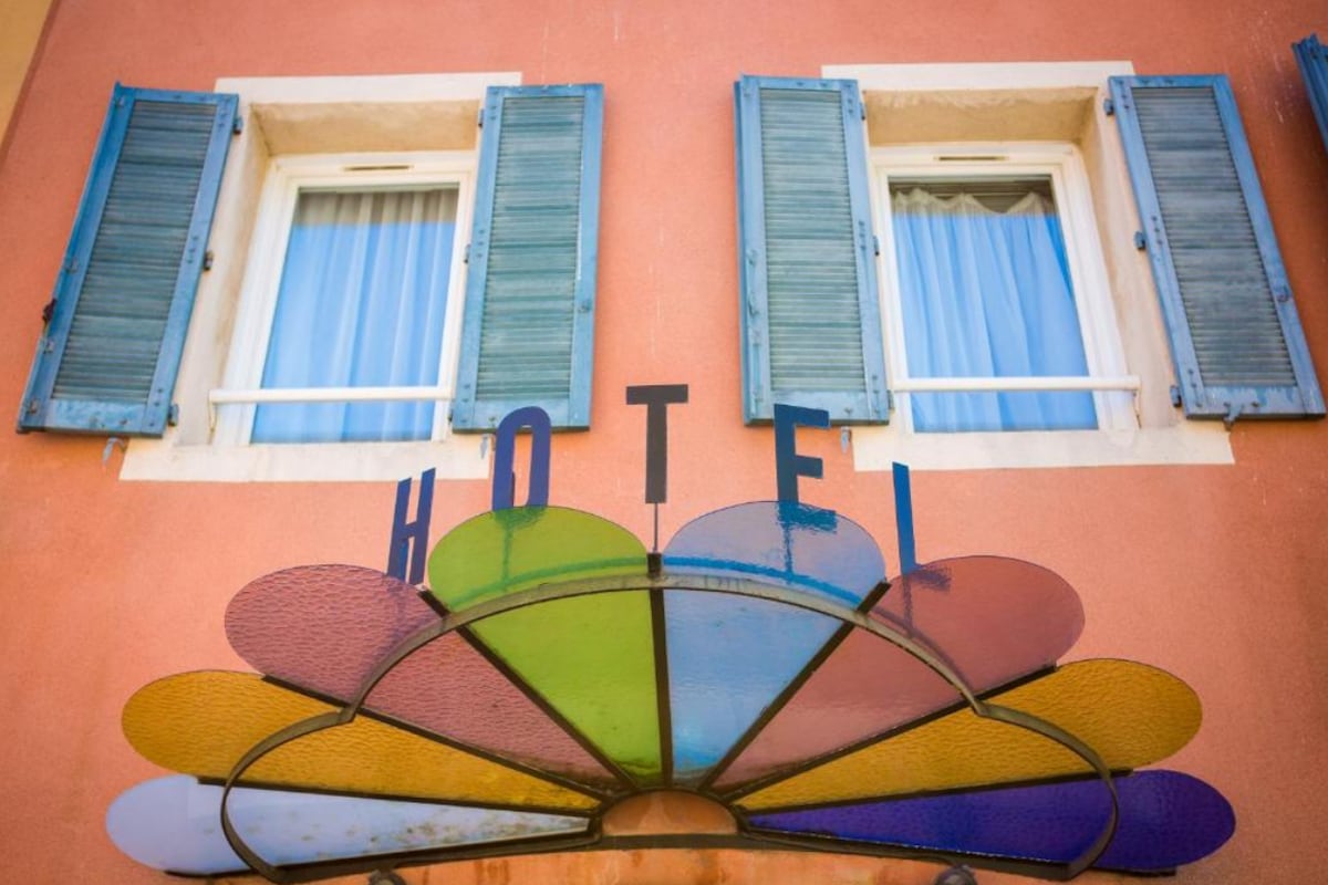 The building features a Mediterranean façade with colorful stained glass above the entrance, reading 'HOTEL.' Two large, blue-shuttered windows are framed by sheer white curtains, adding a charming touch to the architecture.