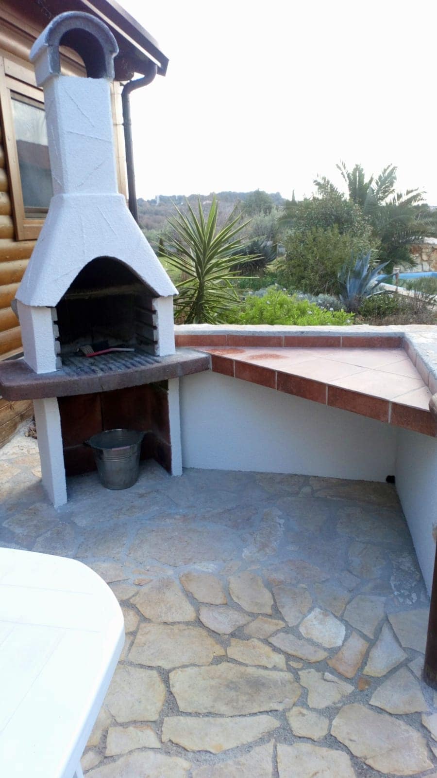 An outdoor barbecue area is featured, showcasing a stone-covered grill with a chimney. The surrounding stone floor complements the rustic aesthetic, while a small bucket sits near the base. Lush greenery and palm trees create a natural backdrop, adding to the tranquil outdoor setting.