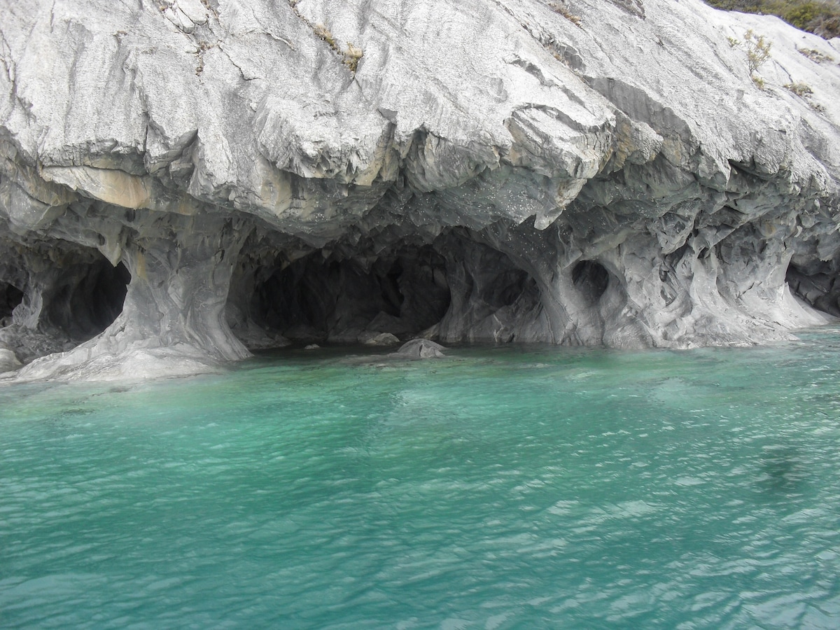 The image captures a dramatic rock formation featuring a series of caves along a shoreline. Clear turquoise water gently flows in front of the textured gray cliffs, highlighting the rugged landscape and unique geological features.