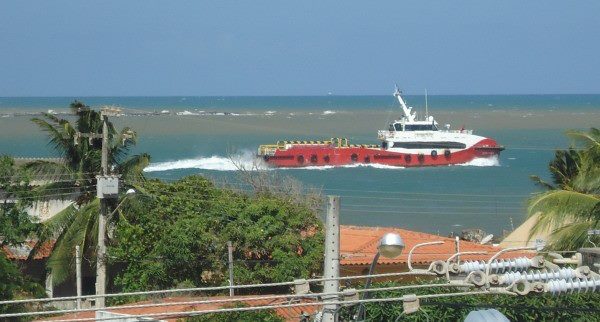 A vessel navigates through the azure waters, creating white waves as it moves. Lush palm trees and colorful rooftops are visible in the foreground, with a clear blue sky serving as a backdrop. The scene captures a moment of coastal activity.