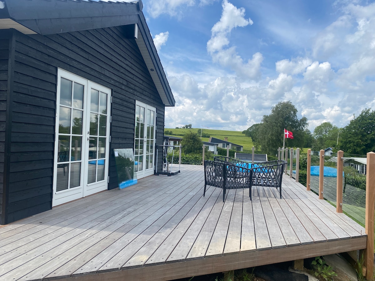 A spacious wooden terrace features a black wicker seating arrangement, positioned for enjoying the surrounding greenery. Large glass doors provide access to the interior, while views of rolling hills are visible in the background under a partly cloudy sky.