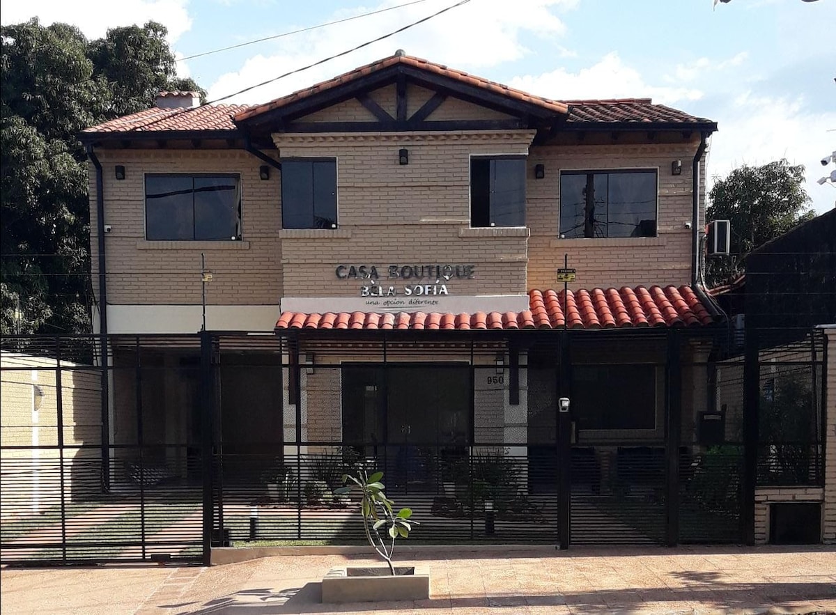 A two-story exterior of Casa Boutique Bèla Sofía is shown, featuring a warm brick facade and a tiled roof. Large windows provide ample natural light, while a secure fence surrounds the property. Greenery is visible in the front yard, enhancing the inviting appearance.