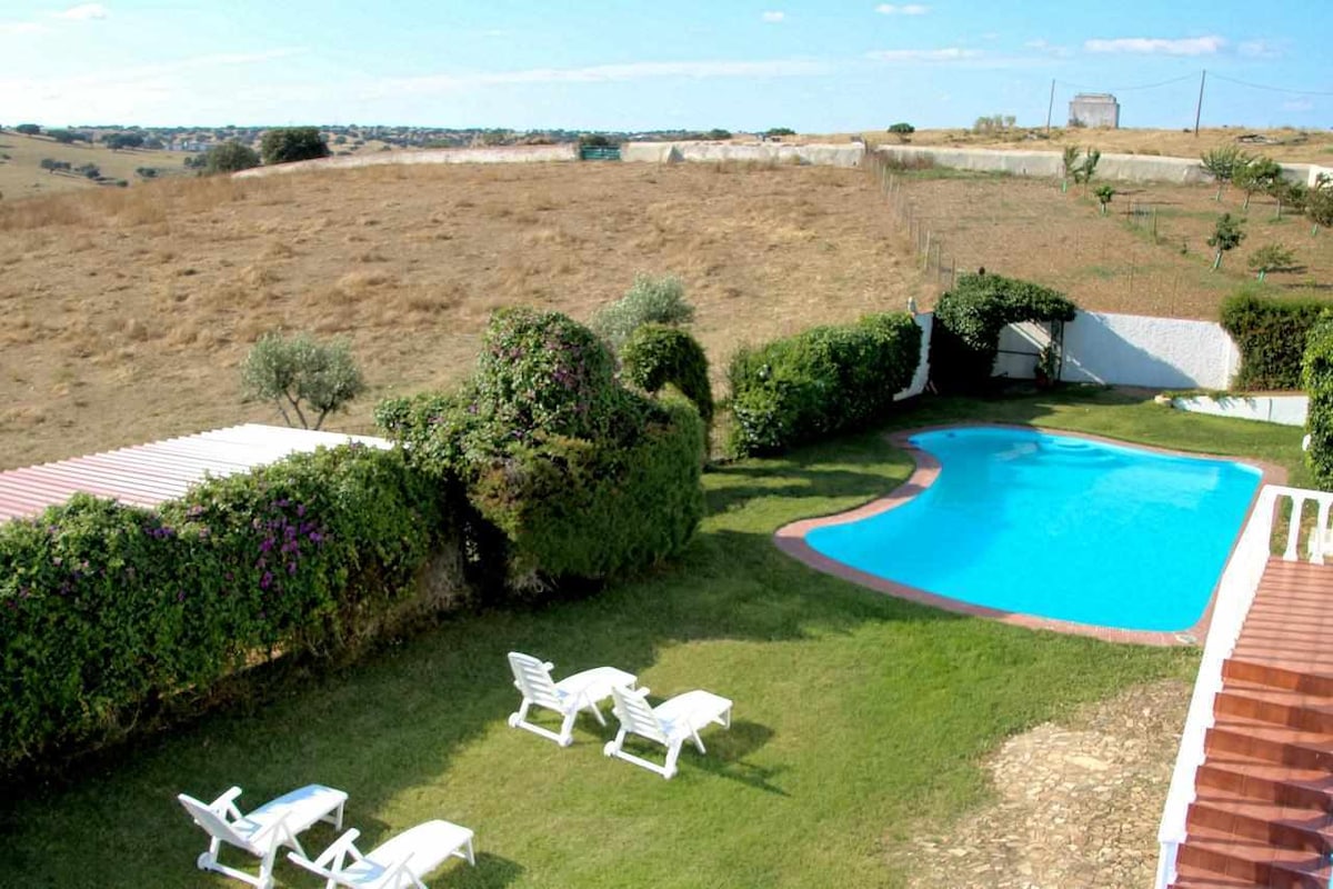 An outdoor area is presented, featuring a clear swimming pool surrounded by lush greenery. Sun loungers are arranged on the grass, offering a relaxed seating option. A gently sloping landscape with distant hills is visible in the background.