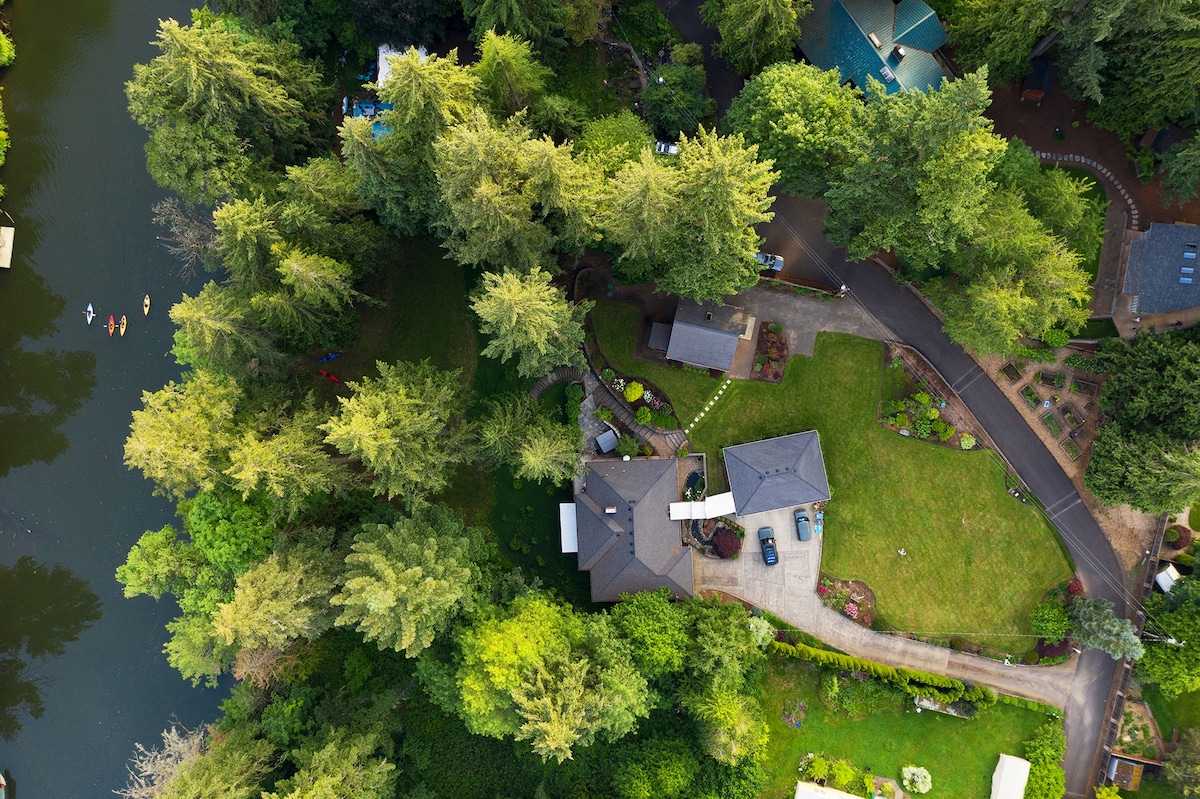 An aerial view showcases the cottage surrounded by lush greenery and tall trees. The property features a well-maintained lawn and a winding pathway. The Tualatin River is visible nearby, with hints of kayaks on the water and distant structures nestled among the foliage.
