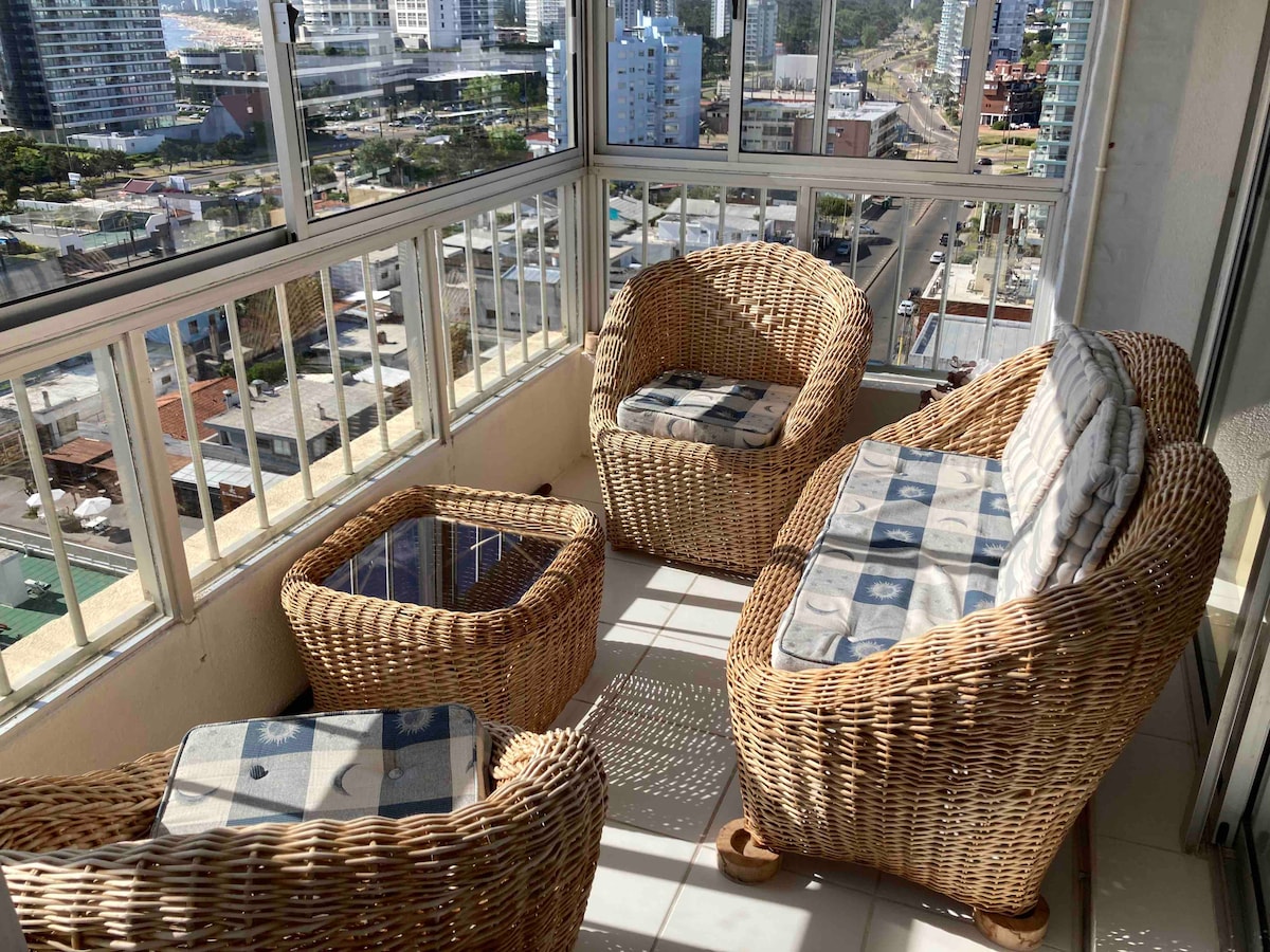 A cozy terrace space features wicker furniture arranged for relaxation, including two armchairs and a sofa. A small table, also wicker, sits at the center. Large windows provide expansive views of the surrounding cityscape, with natural light brightening the area.