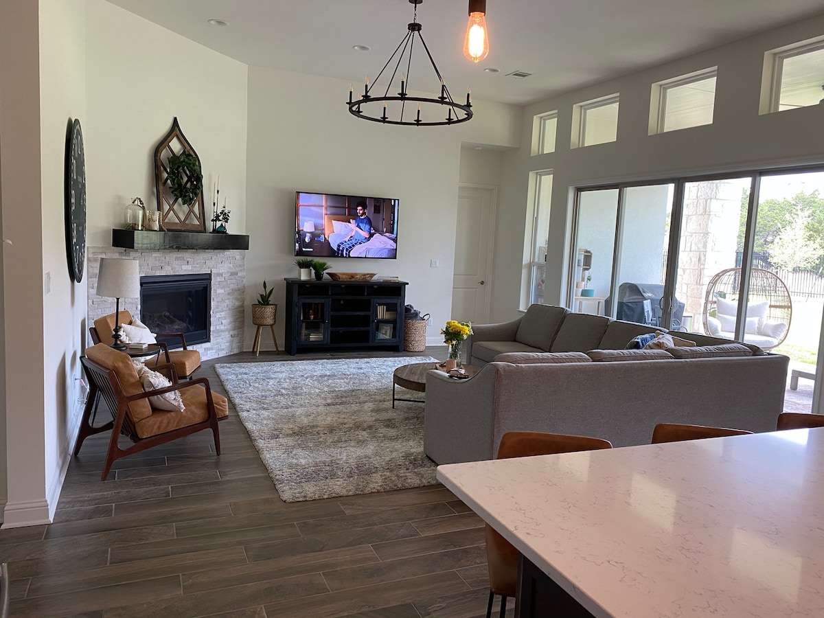 An open-concept living area features a comfortable sectional sofa and two armchairs positioned near a coffee table. A large TV is mounted above a modern fireplace, while light streams in through expansive windows that lead to the outdoor patio. A light-colored rug covers the floor.