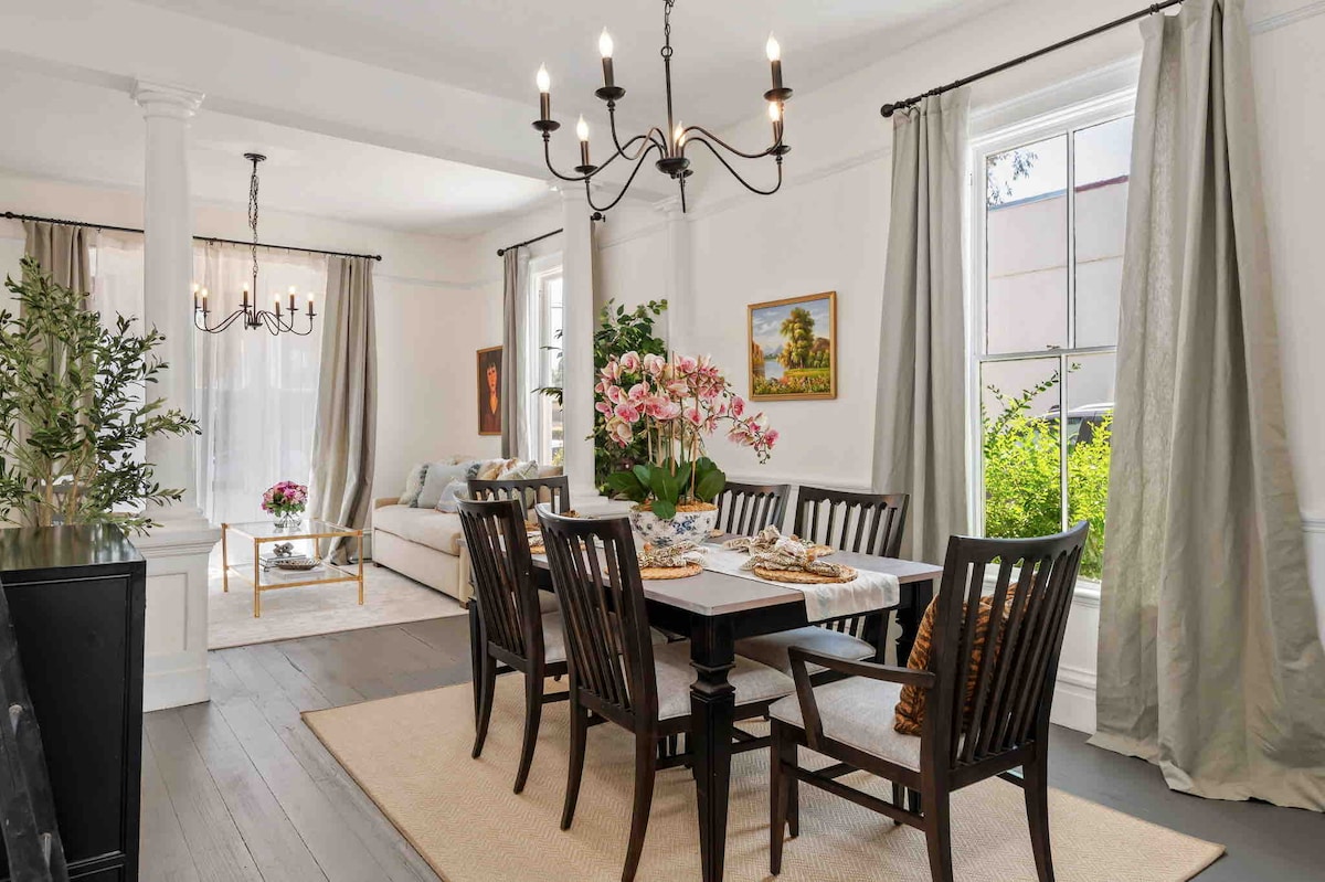 A bright dining area features a dark wood table surrounded by elegant chairs, with a floral centerpiece adding a touch of color. Natural light enters through large windows, while soft drapes frame the view of greenery outside. Adjacent seating offers a cozy atmosphere.