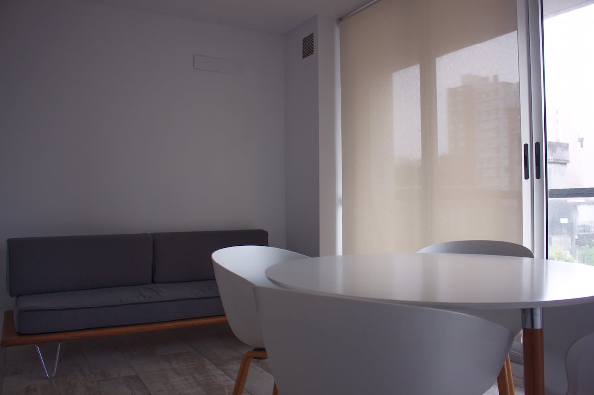 A minimalist living area features a sleek gray sofa positioned against a light-colored wall. A round dining table with white chairs is situated in the center. Natural light filters through large windows covered by sheer shades, creating a bright and airy ambiance.