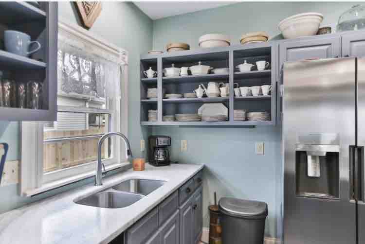 A well-equipped kitchen features light blue walls and white countertops. The cabinetry displays an array of dishes and kitchenware. A double sink is positioned beneath a window, and modern appliances, including a refrigerator and coffee maker, are also visible.