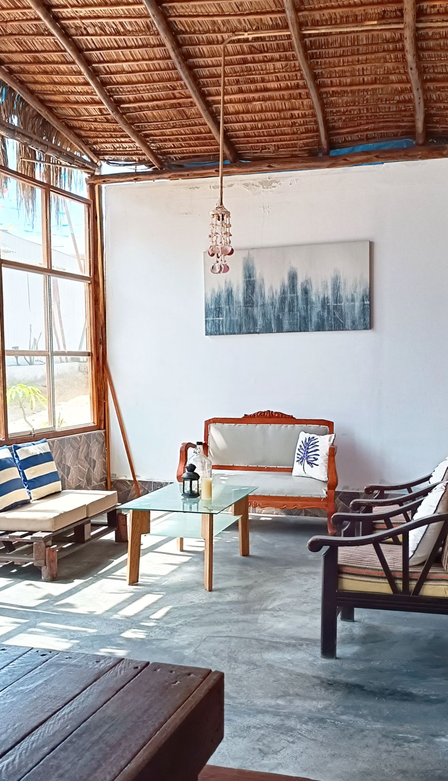 A cozy seating area showcases a mix of wooden and upholstered furniture, with a glass coffee table in the center. A piece of artwork adorns the wall, and natural light floods the space through large windows, creating a bright and inviting atmosphere.