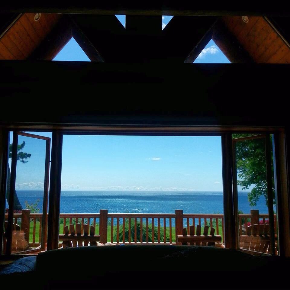 A panoramic view of Lake Michigan is framed by large open glass doors. The spacious exterior deck features wooden railings and overlooks the serene water with distant clouds visible against a clear blue sky. The natural light floods the interior space.