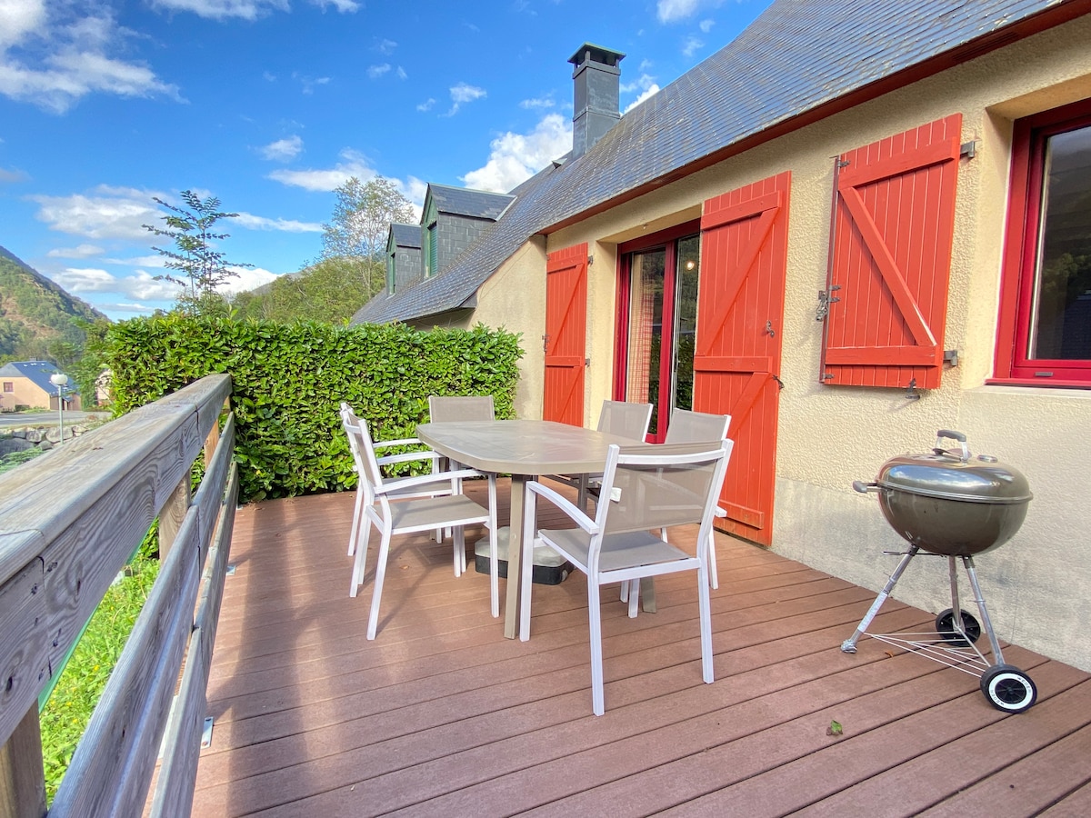 A terrace is showcased, featuring a dining table with four chairs and a barbecue grill. Surrounding greenery and the red shuttered doors add a welcoming touch. The scenery includes distant mountains under a clear sky, promoting outdoor relaxation and dining.