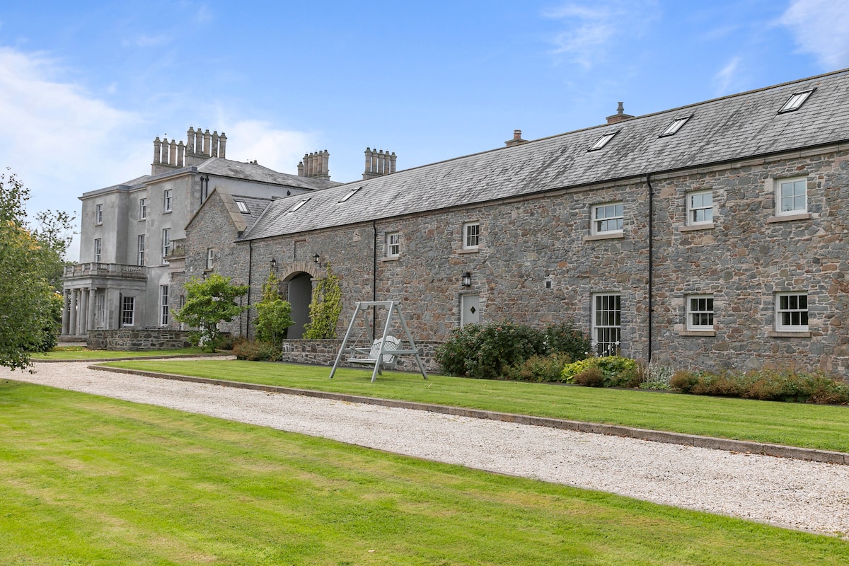 The exterior of Florida Manor is showcased, highlighting the original stone buildings surrounded by expansive landscaped lawns. A swing set is visible on the grass, with a gravel path leading towards the entrance of the manor, framed by trees and a clear blue sky.