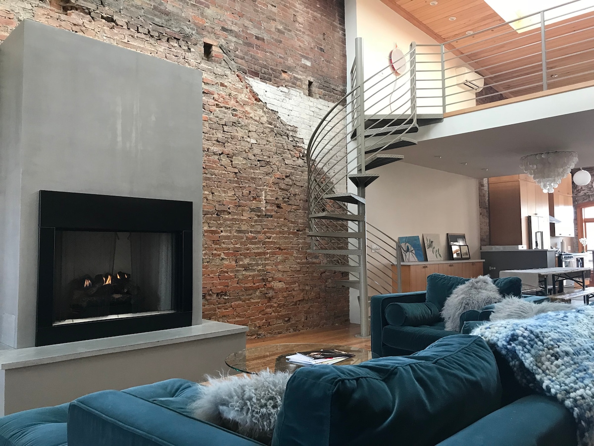 A spacious living area features a modern gas fireplace set against a rustic brick wall. A curved staircase leads to the upper level, while plush turquoise sofas adorned with cozy throws invite relaxation. Natural light floods the room, enhancing its open atmosphere.