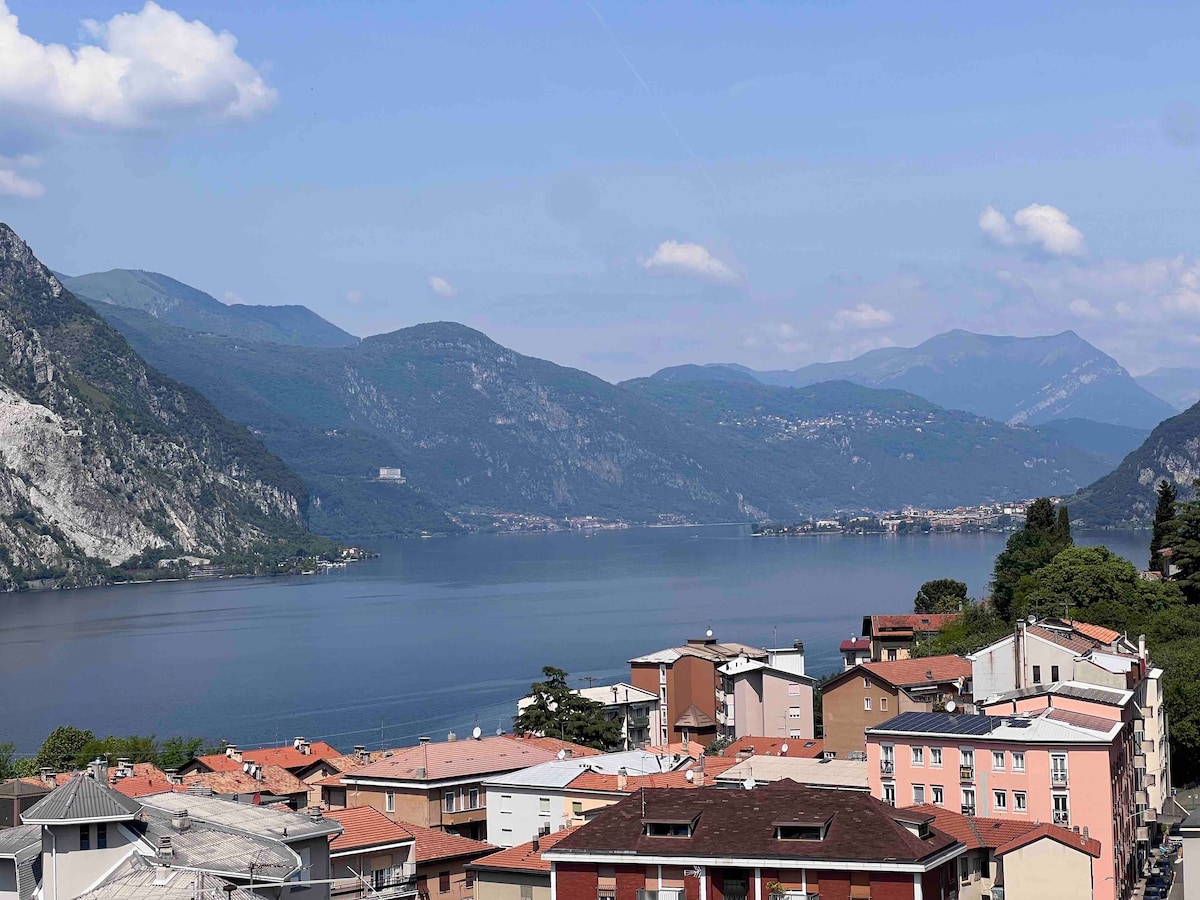 A serene view of the lake and surrounding mountains is captured, showcasing calm water reflecting the sky. Residential buildings with varied architectural styles are visible in the foreground, while lush green hills rise in the background under a clear blue sky.
