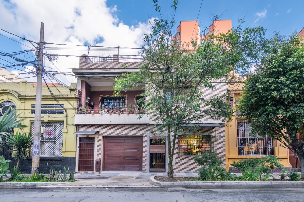 A charming building features a unique brick facade with a harmonious blend of colors. Lush greenery surrounds the structure, enhancing the inviting appearance. A wooden garage door and potted plants adorn the entrance, while windows are flanked by vibrant flower boxes, adding character to the exterior.