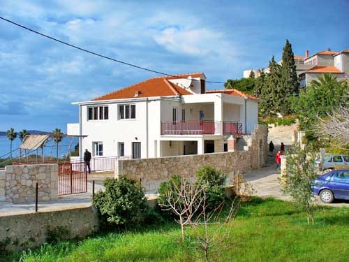 A spacious family house is depicted, featuring a bright white exterior and a red-tiled roof. The property includes a balcony overlooking a green area and the sea. A driveway leads to the house, bordered by trees and plants, creating a calming environment.