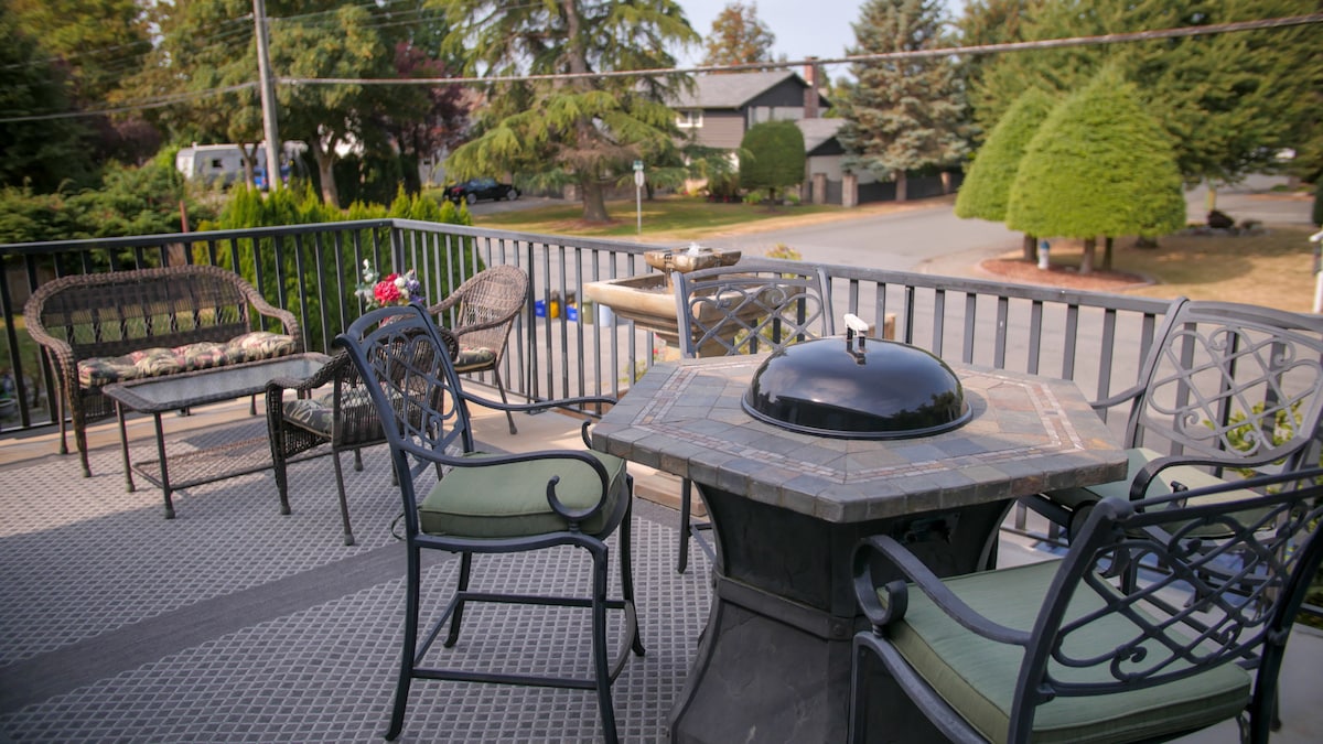 An outdoor deck is furnished with metal and fabric seating, offering a comfortable space for relaxation. A stone-topped table with a grill sits at the center. Lush greenery and a view of the neighborhood can be seen in the background.