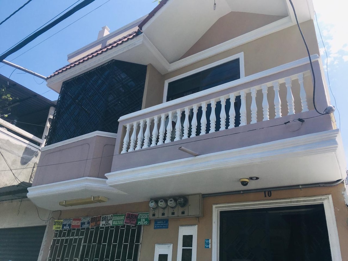 A three-story peach-colored home is depicted, featuring a second-floor balcony with white balustrades. The entrance showcases glass doors, adorned with various utility meters. A sunny blue sky can be seen above, reflecting a bright and open atmosphere.