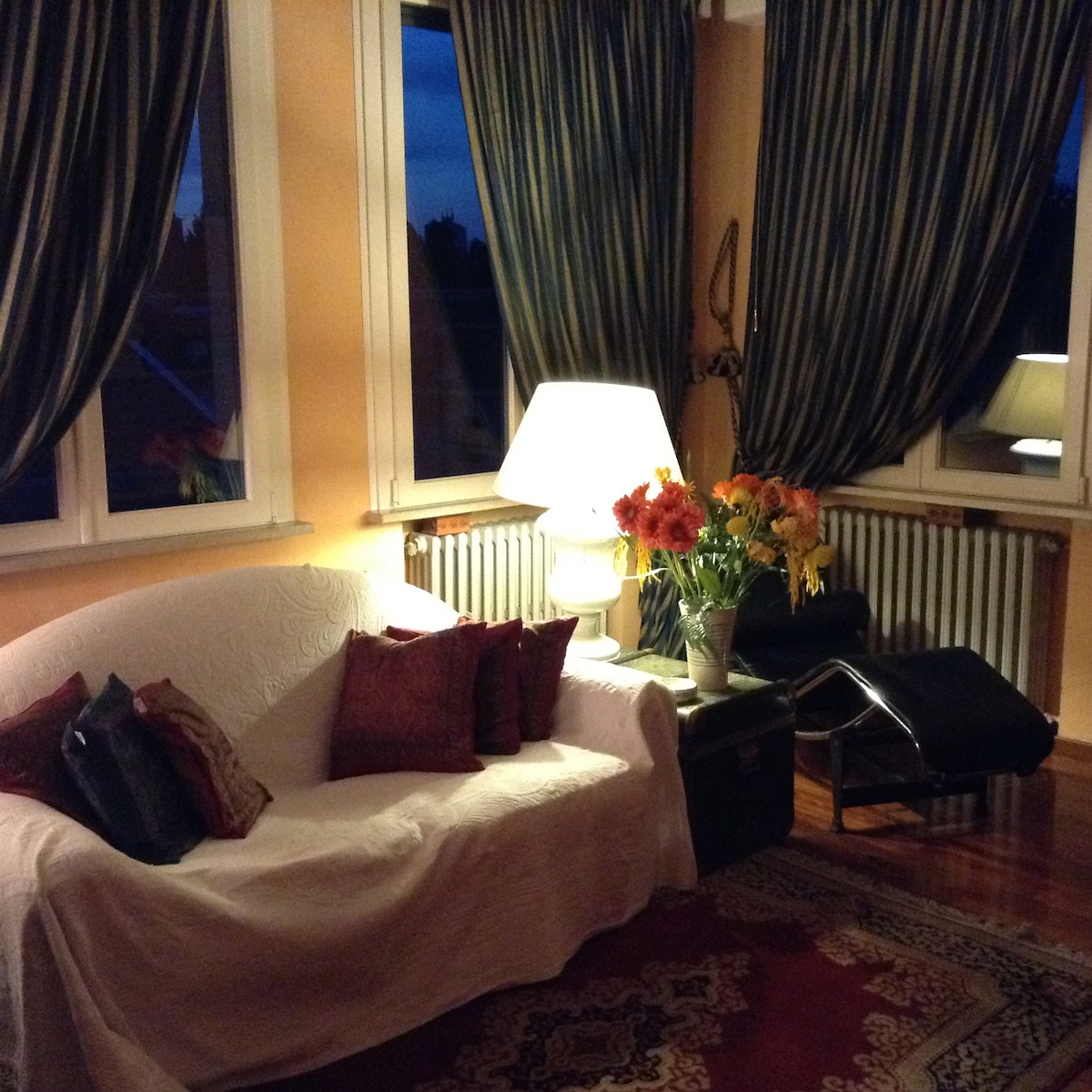 A spacious seating area features a white couch adorned with colorful cushions. A stylish floor lamp illuminates the space, complemented by a decorative vase of vibrant flowers. Dark curtains frame large windows that provide views of the outside, enhancing the cozy atmosphere.