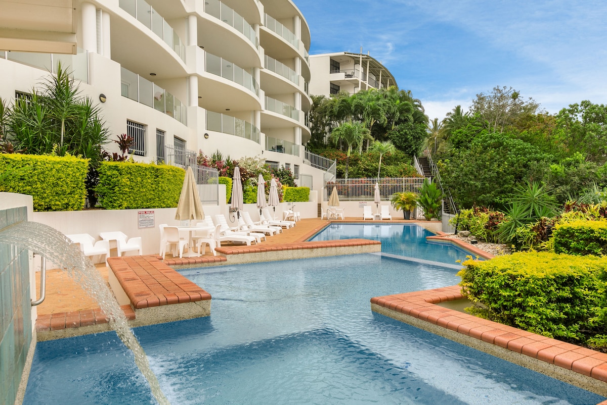 The resort pool area features a shimmering blue pool surrounded by landscaped gardens. Several sun loungers are positioned along the pool's edge, with shaded areas provided by umbrellas. Fountains gently flow into the pool, creating a serene atmosphere and enhancing the tropical setting.