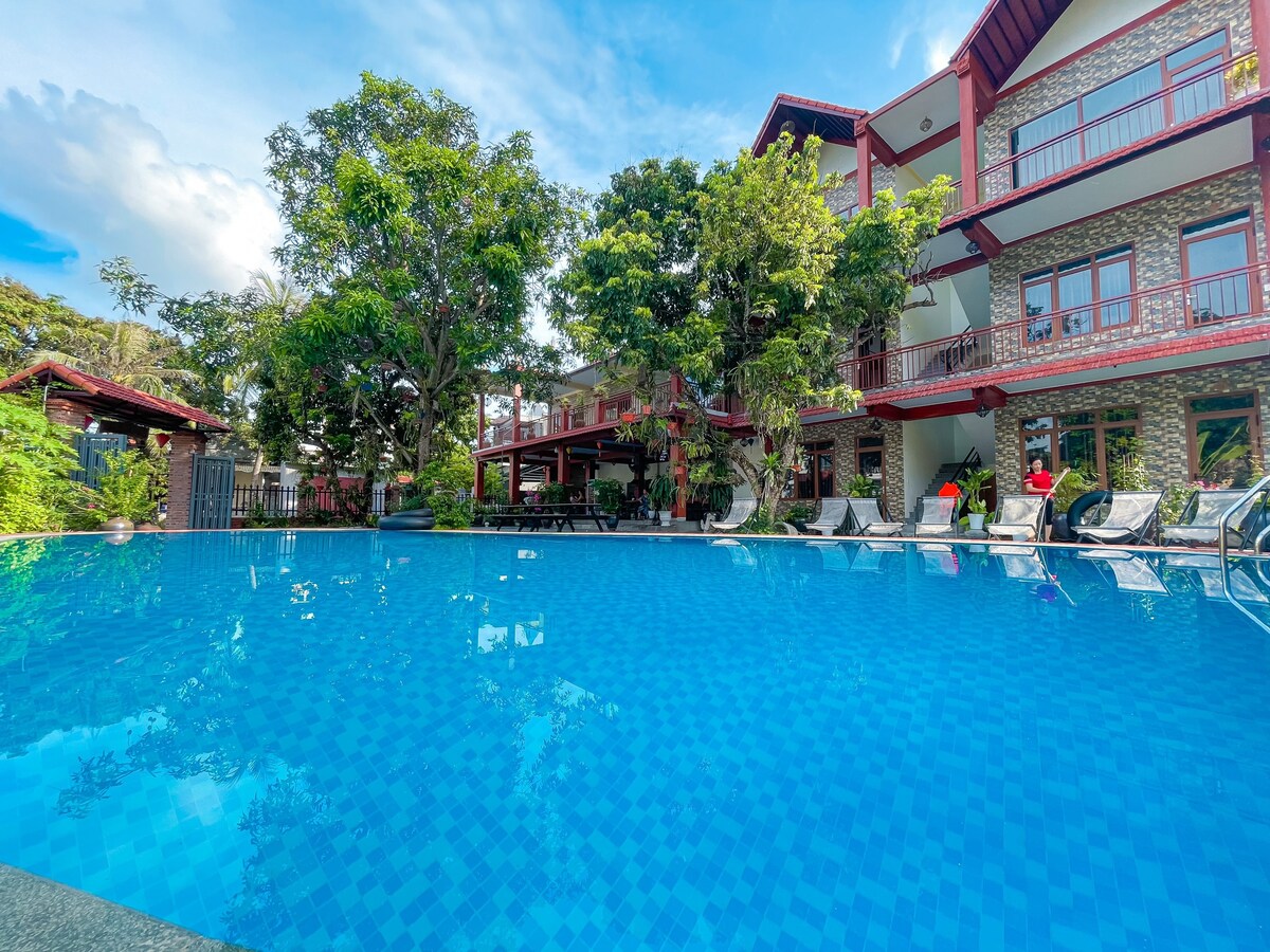 A large swimming pool is surrounded by greenery and complemented by a stone facade building. Sun loungers are positioned around the pool, inviting relaxation. Balconies on the upper levels overlook the pool area, enhancing the spacious atmosphere.